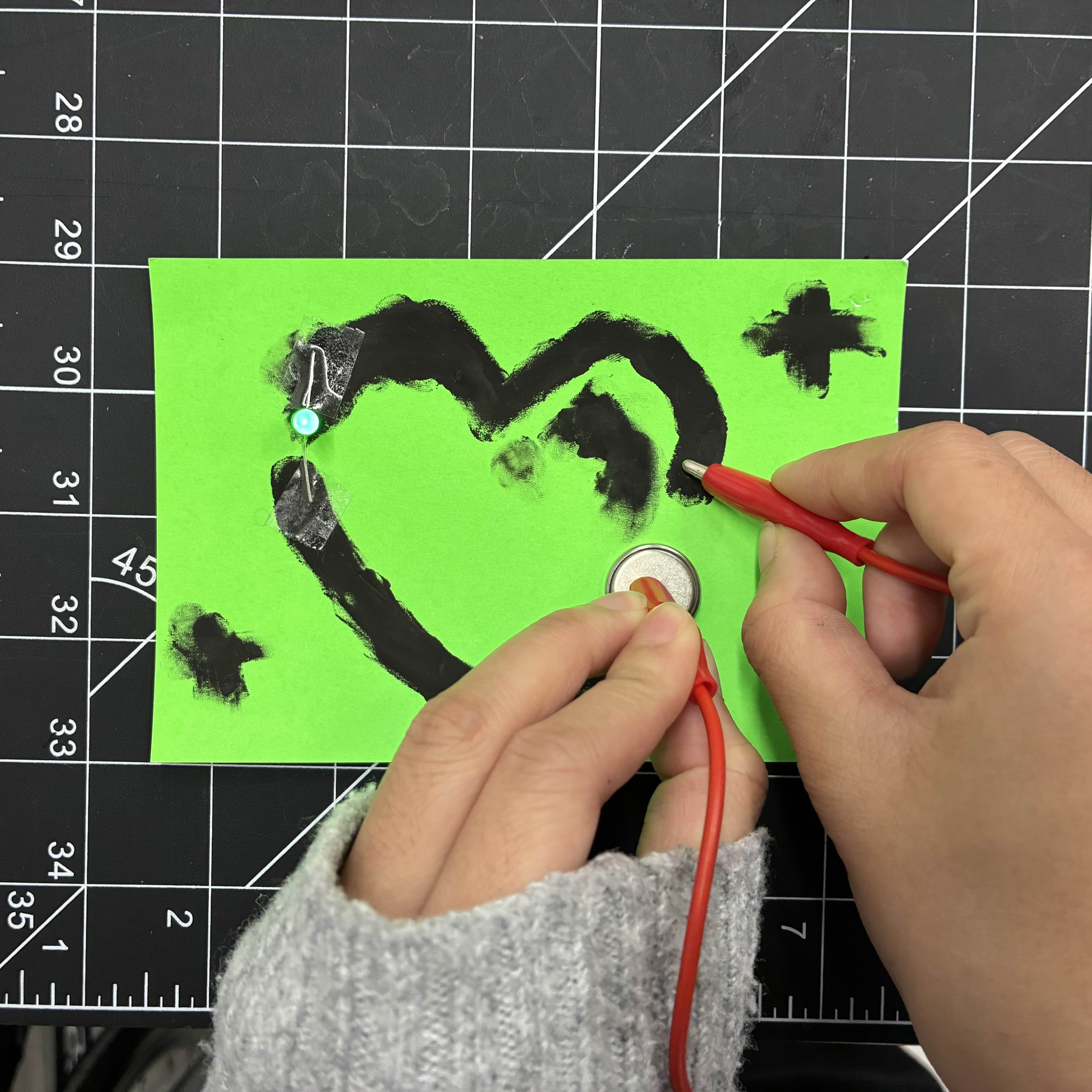 This is the heart made of conductive paint from a higher angle. I used a green light to match the green paper that I used. I taped an LED light to fill the gap in the heart and then placed a battery on the other end. I then used an alligator clip wire to connect these items together so the light can glow.
