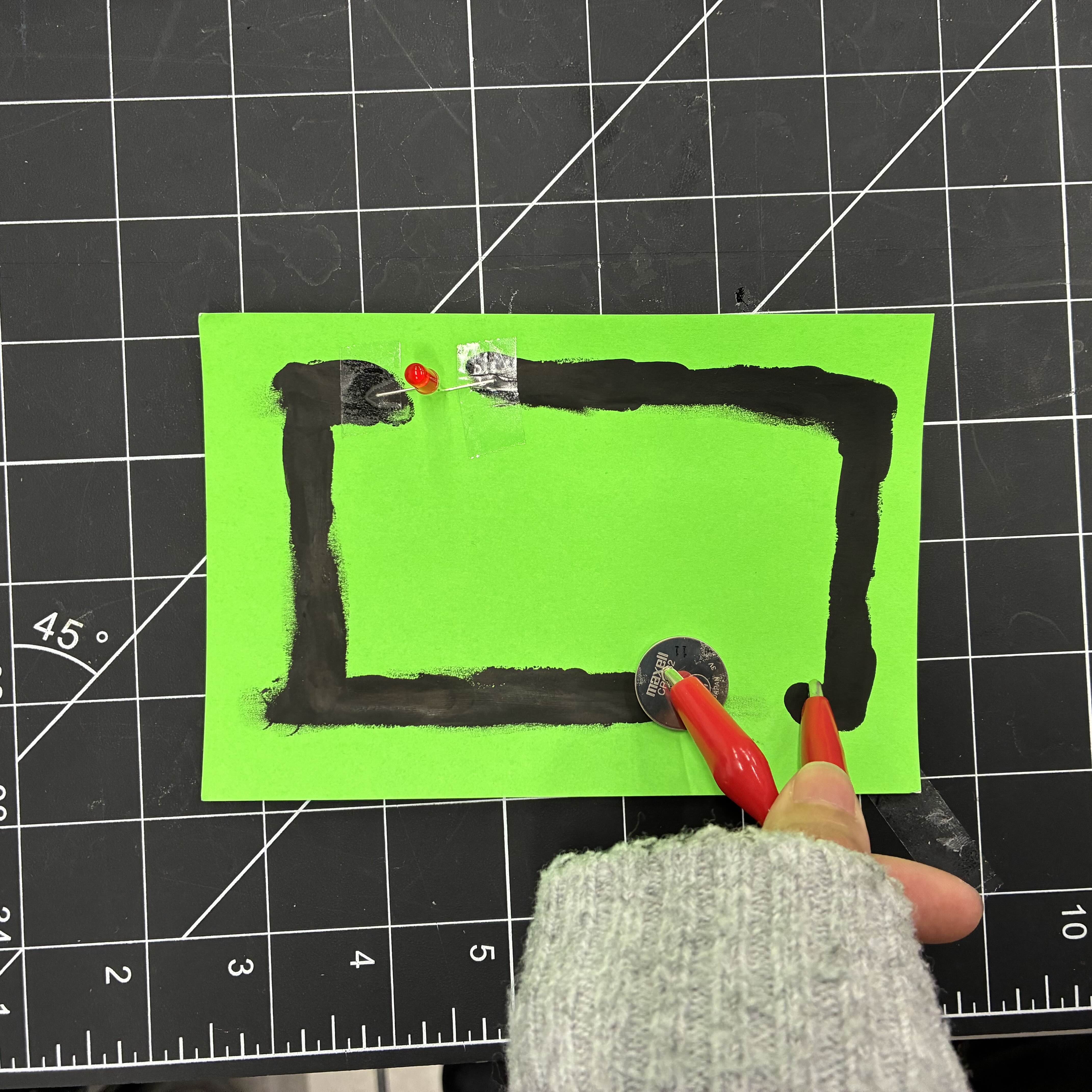 This is the rectangle made of conductive paint. I taped a red LED light to contrast the green and to fill the gap in the rectangle and then placed a battery on the other end. I used an alligator clip wire to connect these ends to make the light glow.